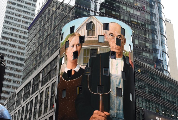 "American Gothic" appears on a tall, rounded building, its windows creating dark gaps in the scene of a woman and man standing in front of their house, the pitchfork in his hand between them. The building is surrounded by taller buildings. 