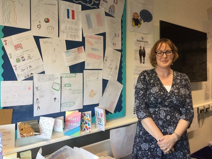 Charlotte Nash stands with her hands in front of her and wears glasses and a black and blue floral dress. She stands in front of a bulletin board displaying student's work and thank you notes.