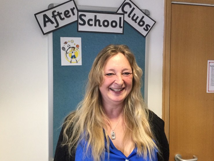 Clare Selby has long blonde hair and smiles toward the camera in front of a board that advertises after school club activities.