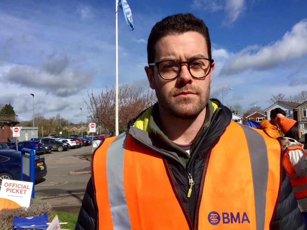 Dr. Michael McArdle has brown hair and a beard. He wears black-framed glasses and an orange vest and looks solemnly at the camera.