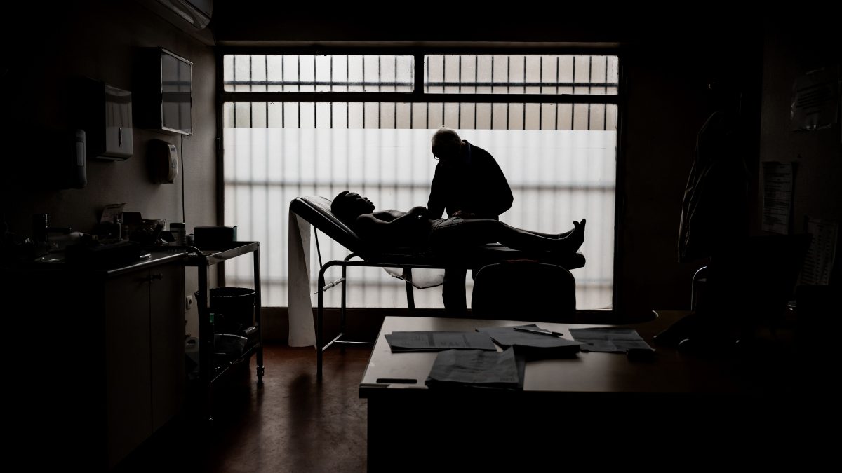 A doctor examines a patient in a clinic. Because of the backlight, both the patient and the doctor can be seen almost in silhouette.