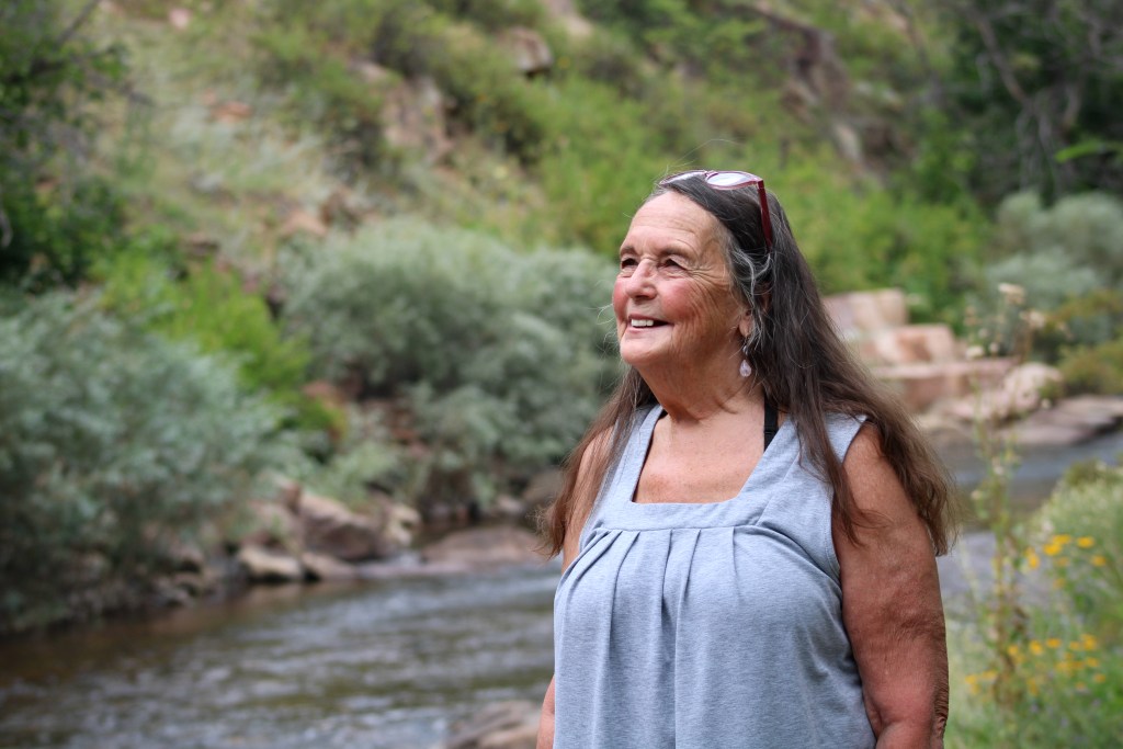 Une femme vêtue d’un chemisier bleu clair regarde au loin en souriant.  Elle se tient devant un ruisseau.