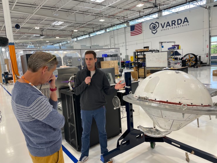 Varda CEO Will Bruey stands inside the warehouse, showing the white Varda reentry capsule to Marketplace's Kai Ryssdal. 