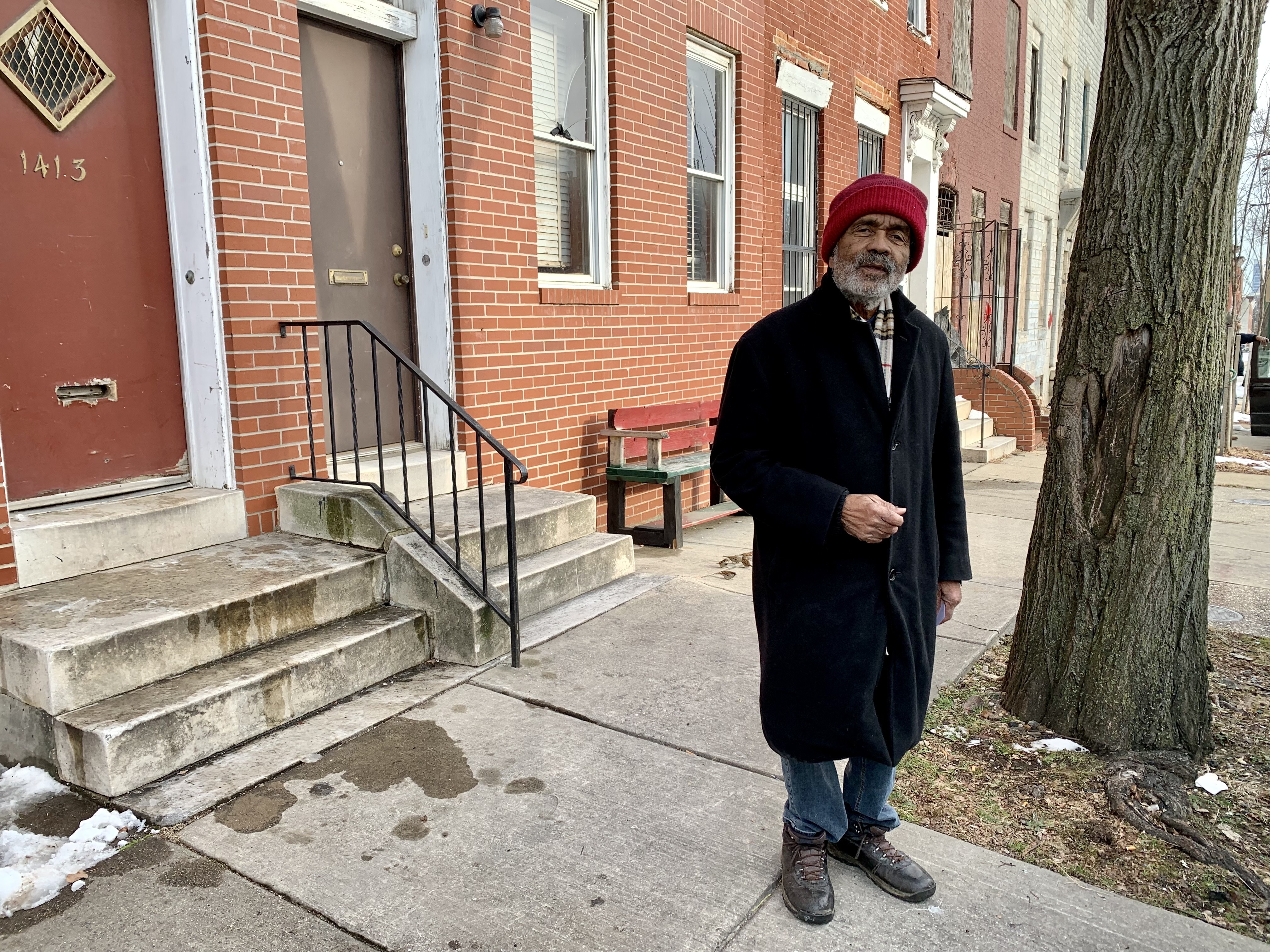 Charles Dugger, a Black man wearing a black coat and a red hat, stands on the sidewalk in front of several rowhouses