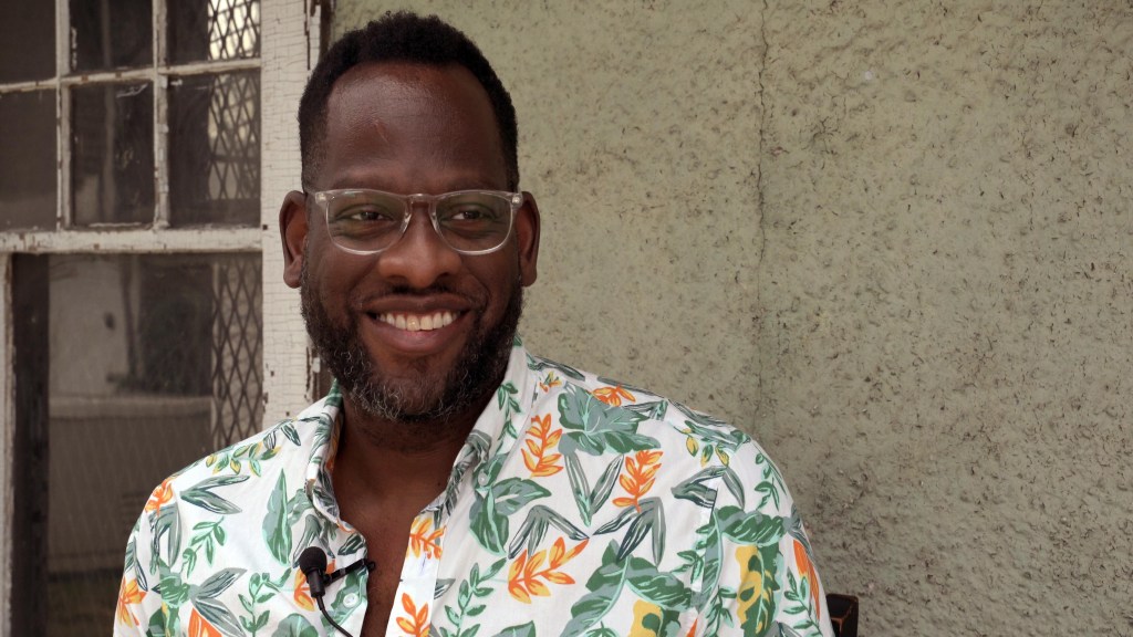 Evan Narcisse wears clear glasses and a floral shirt. he is smiling 