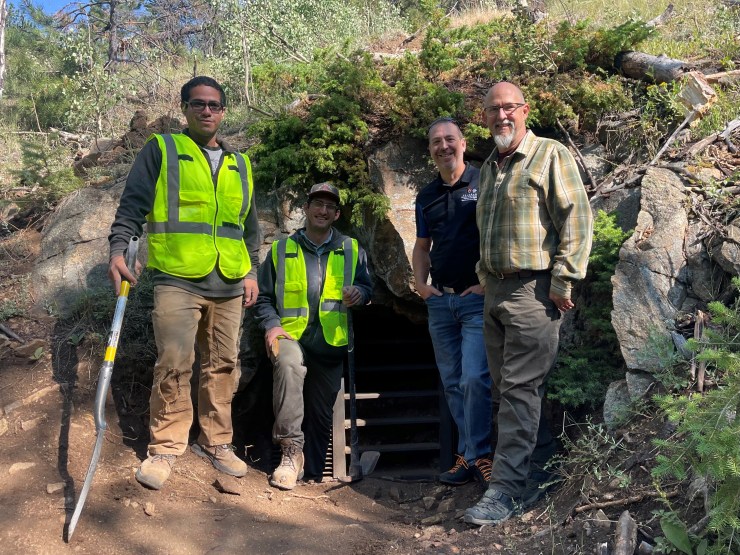 Four men stand in front of an abandoned mine. Two wear yellow safety vests.