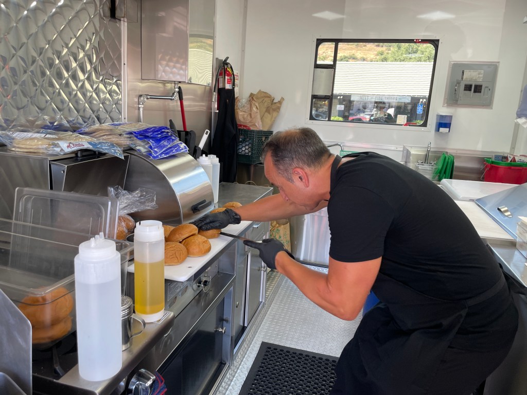 In a narrow galley kitchen, a man dressed in black bends down to cut hamburger buns in half.