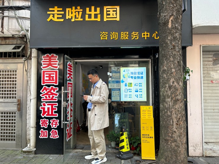 An Asian man wearing a brown trench coat and khakis looks at his phone and stands in the doorway of a store.