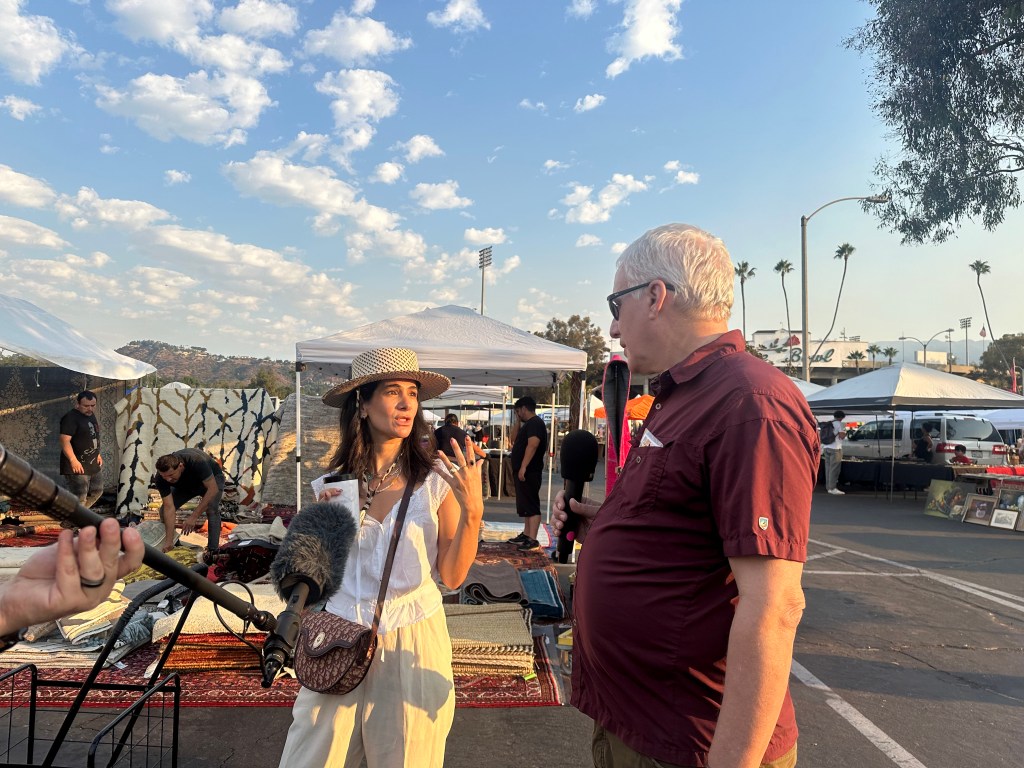 A person holds a mic toward a man and a woman talking. The woman, center, is wearing a white top and cream-colored pants and sunhat. The man is older, with gray hair and a maroon shirt. They're standing in a parking lot turned into a flea market, where vendors are displaying rugs and art behind them. The sky is partly cloudy. 