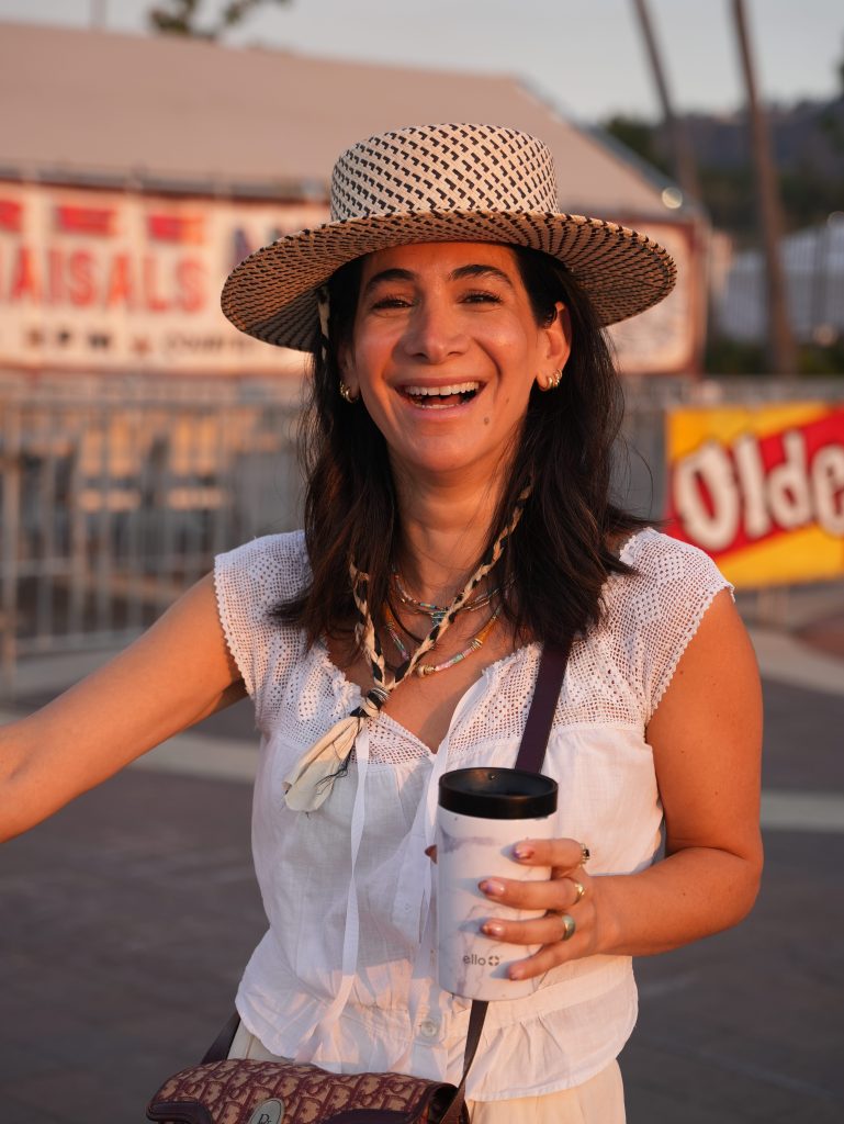 A woman with black hair in a white shirt and sunhat holds a coffee and smiles brightly.