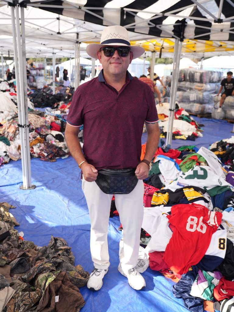 A man in sunglasses, a maroon shirt, white pants, with a light-colored sunhat and black fanny pack stands under a striped tent. The floor is blue and there are piles of shirts and jerseys around him.