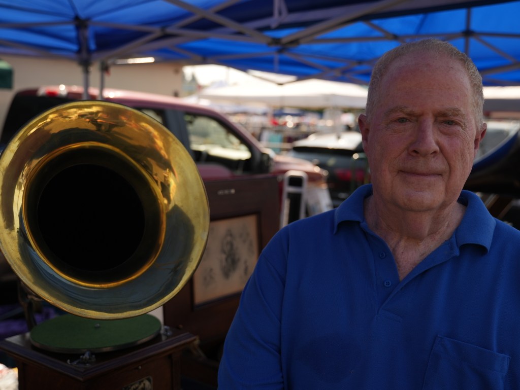 An older bald man in a blue shirt stands near a brass-colored phonograph.