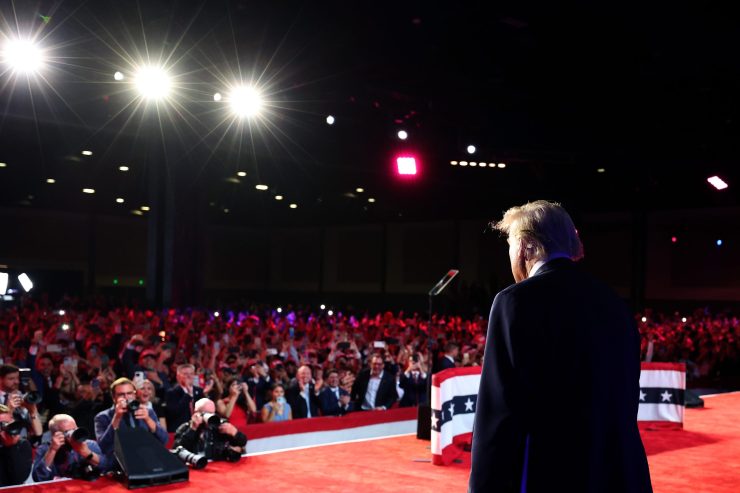 The back of former President Donald Trump as he steps onto a red stage in front of a sea of ​​people.