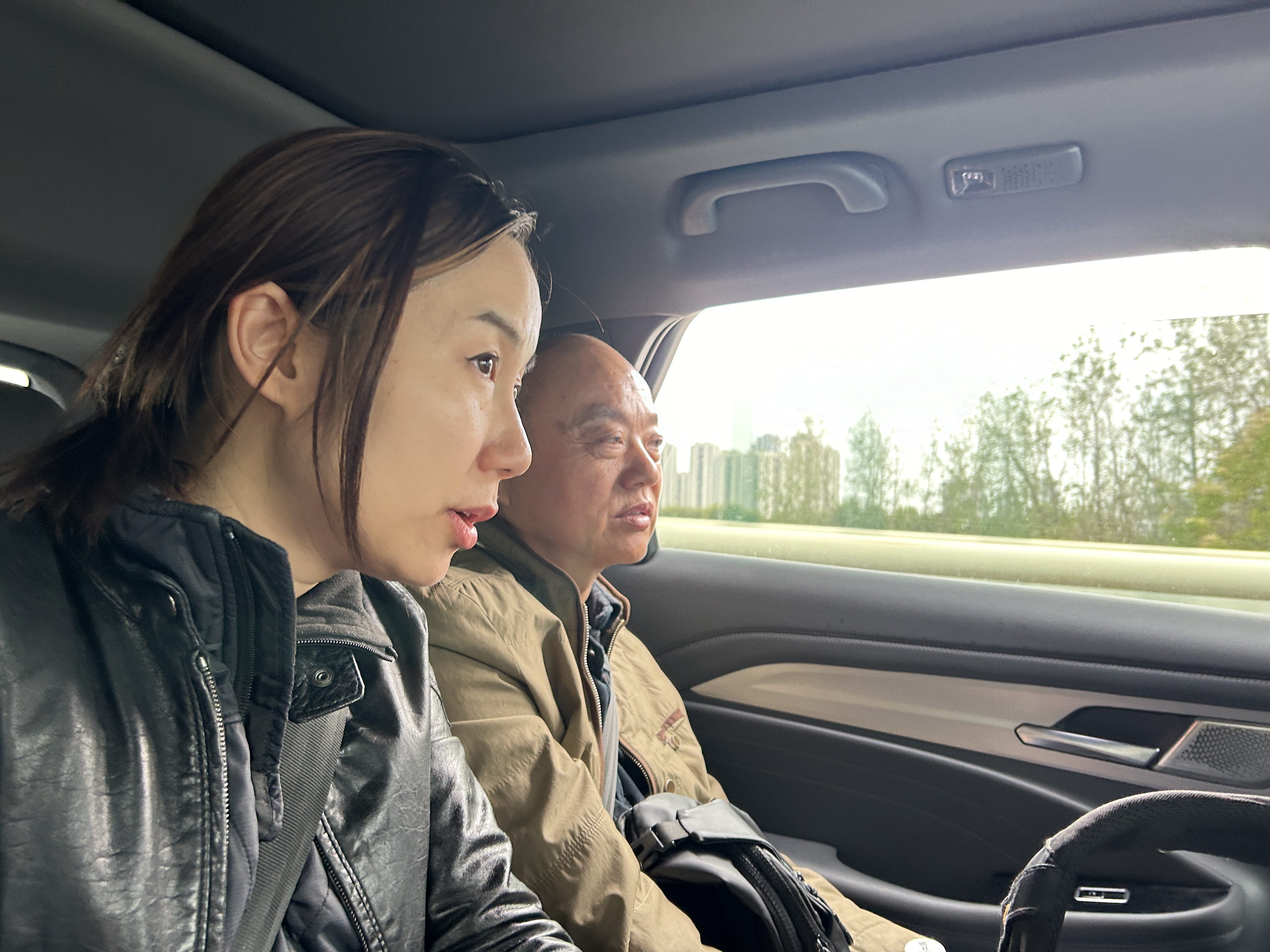 An Asian man and woman wearing seat belts are sitting in the back of a car. The man is sitting next to a window with a view of trees and tall buildings. 