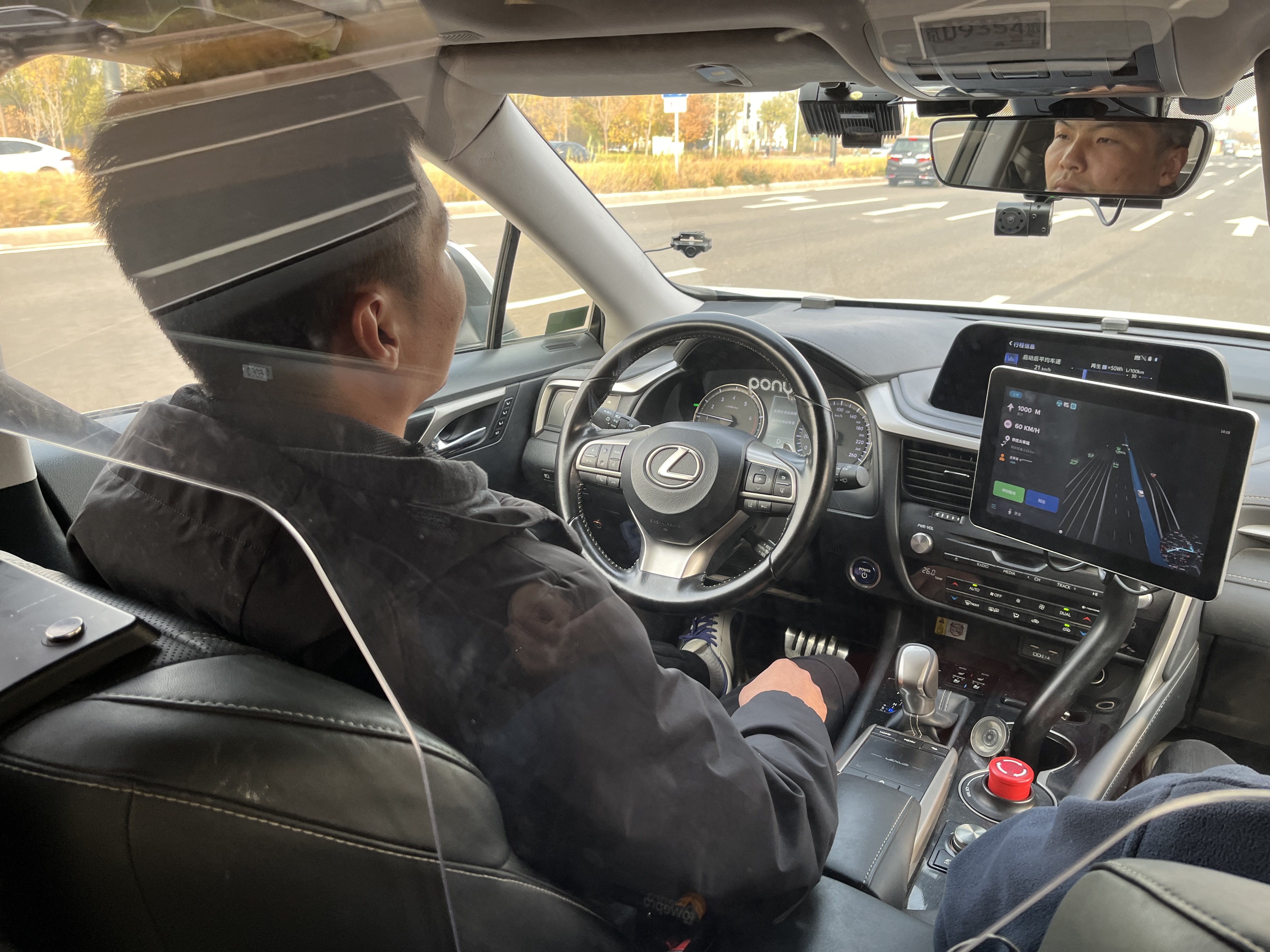 A driver, seen from the back seat and through a transparent partition, sits in a car with his hands at his sides. The car is on a road, the image of which is displayed on a screen next to the steering wheel.