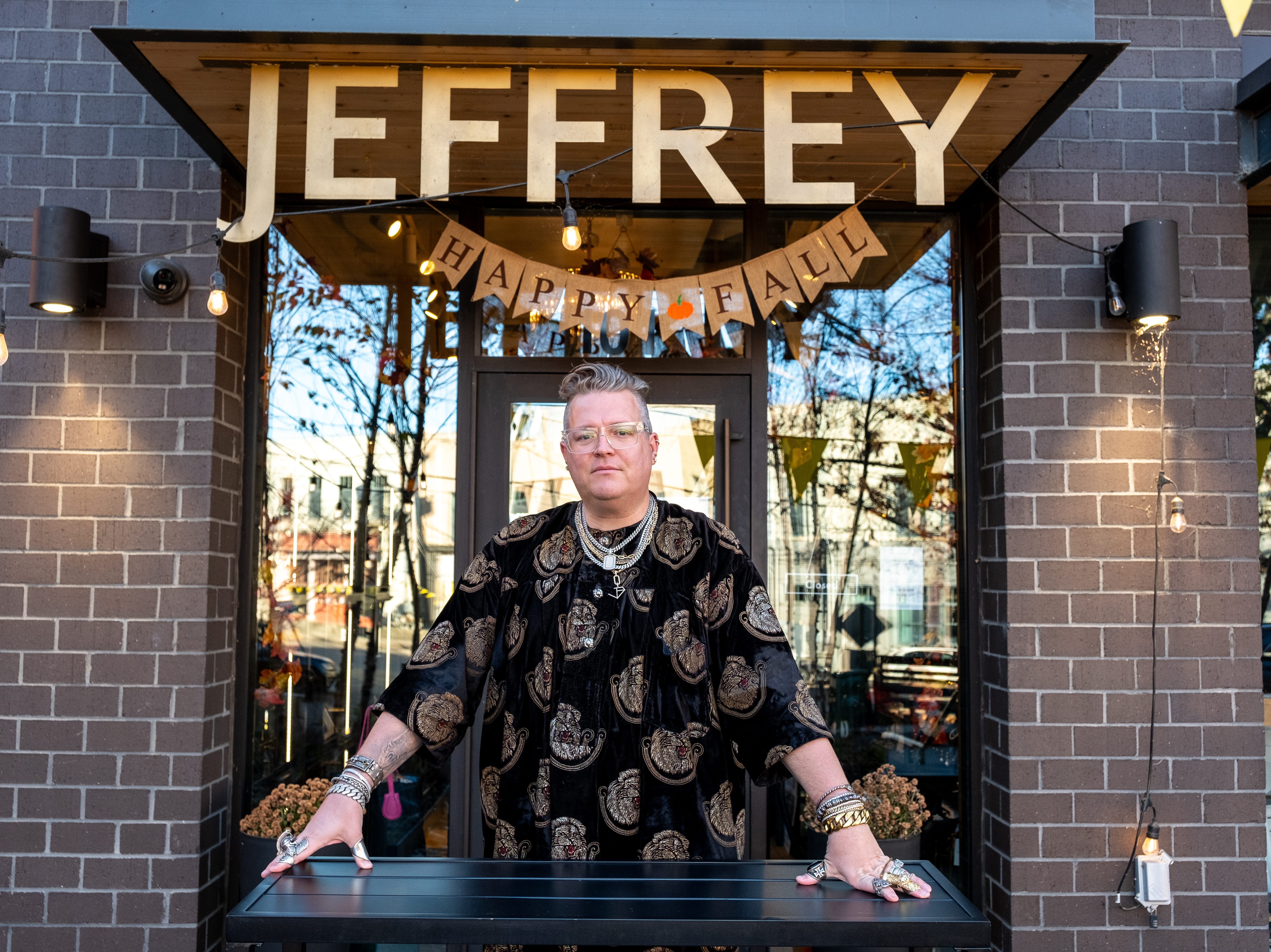 A person who wears a lot of silver jewelry and a black patterned shirt. They are standing in front of a small storefront. A sign that says "Jeffrey" in large letters, hangs from the awning with a "happy autumn" sign underneath.