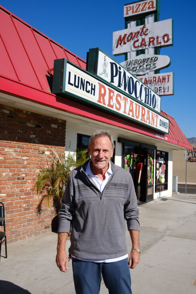 A man in a gray zip-up stands outside a one-story brick building with vintage signage advertising the Monte Carlo Italian Deli and Pinocchio restaurant.