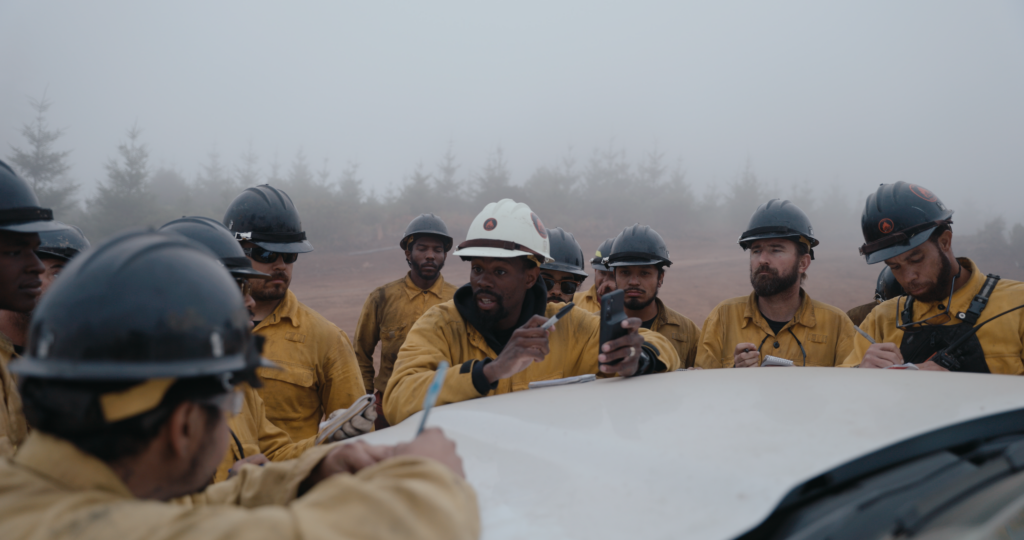 A group of men in yellow jackets and hardhats gathered around the hood of a vehicle. Some take notes on pads of paper.