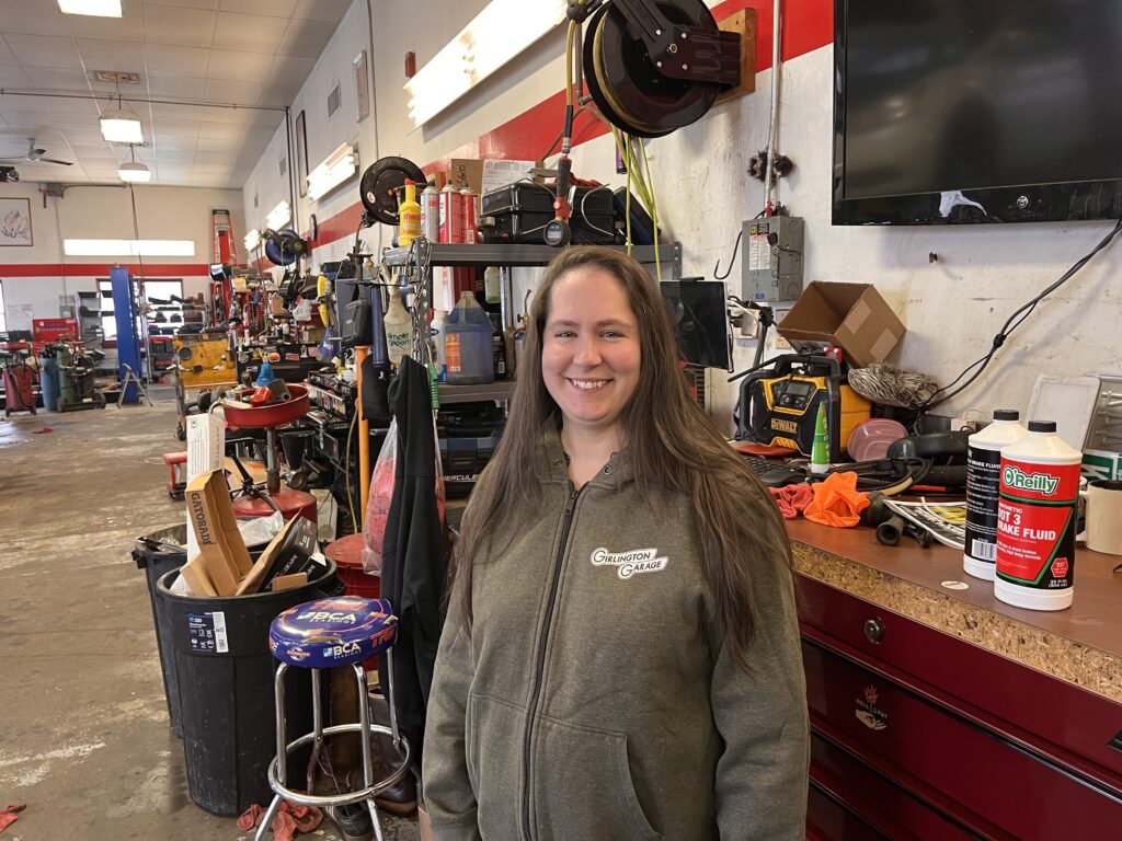 A woman in a grey hoodie stands in front of a workbench in an auto repair shop.